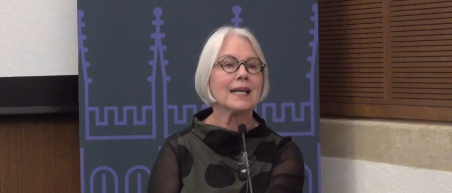 Rosemarie Garland-Thomson stands at a podium at Oxford University. She has silver hair, black spectacles, and is wearing a brown blouse with black polka dots.